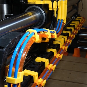 May include: Close-up of a machine with yellow, blue, and orange tubes and wires. The tubes are held in place by yellow brackets. The machine has black and silver components. The background is a wooden surface.