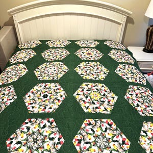 May include: A green quilt with a repeating pattern of white and floral hexagon shapes. The hexagons are filled with a colorful design of flowers and birds. The quilt is on a bed with a white headboard.