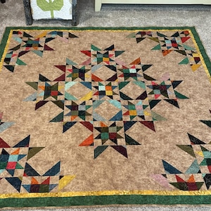May include: A square quilt featuring a central star pattern composed of multi-colored fabric patches in shades of red, blue, green, and yellow. The background is a mottled tan fabric, and the quilt is finished with a yellow and green border.