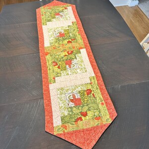 May include: A rectangular table runner with a patchwork design. The runner features a central section with green and cream-colored floral patterns, bordered by a solid orange fabric. The runner is placed on a dark wooden table.