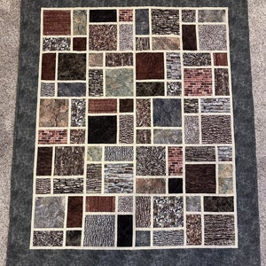 May include: A patchwork quilt featuring a variety of textured fabric squares in shades of brown, gray, and red. The squares are arranged in a grid pattern and separated by off-white strips. The quilt has a dark gray border.