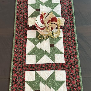 May include: A rectangular quilted table runner featuring four white blocks with green star patterns and a black border with a red poinsettia print. A ceramic bowl shaped like a wrapped candy with red and white swirls holds a string of gold beads.