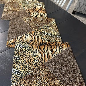 May include: A rectangular table runner featuring a patchwork design of various animal prints including leopard spots and tiger stripes in shades of brown, black, tan, and gold. The fabric is quilted with visible stitching along the edges and seams.