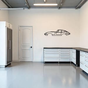 May include: A modern garage interior with white walls and a gray floor. A black metal car silhouette is mounted on the wall. White cabinets with black countertops and a stainless steel refrigerator are also present.