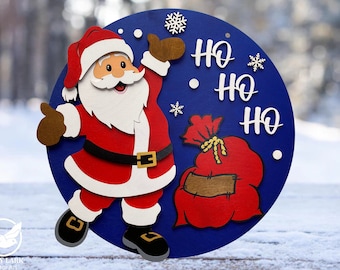 May include: A blue circular wooden sign with a white background. Santa Claus is depicted in a red suit with a white beard and a white trim. He is holding a bag of gifts. The sign says "Ho Ho Ho" in white letters.