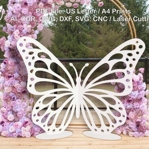Può includere: Una grande decorazione a farfalla bianca tagliata al laser con dettagli intricati sulle ali. La farfalla è posizionata su una superficie di legno, con una composizione floreale di fiori rosa e viola sullo sfondo. Il testo nell'immagine recita "PDF File: US Letter / A4 Prints".