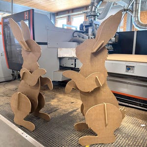 May include: Two standing rabbit figures crafted from light brown medium-density fiberboard. Each rabbit features a flat, cutout silhouette design with upright ears, bent front paws, and a rounded tail, assembled with interlocking slotted pieces.
