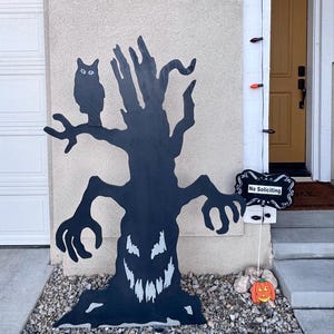 May include: A black wooden cutout of a gnarled tree with a jagged, toothy face and an owl perched on a branch. Next to the tree is a black sign with white decorative borders and the text No Soliciting, plus a small orange jack-o'-lantern.
