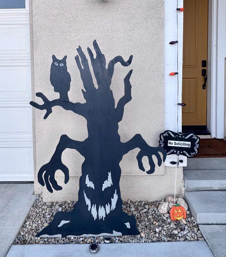 May include: A black wooden cutout of a gnarled tree with a jagged, toothy face and an owl perched on a branch. Next to the tree is a black sign with white decorative borders and the text No Soliciting, plus a small orange jack-o'-lantern.