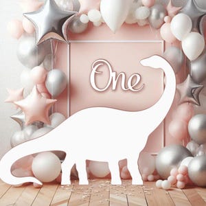 May include: A white dinosaur cutout stands before a pink backdrop with the word "One" in script. The backdrop is decorated with pink, silver, and white balloons, including star-shaped balloons. A party decoration.