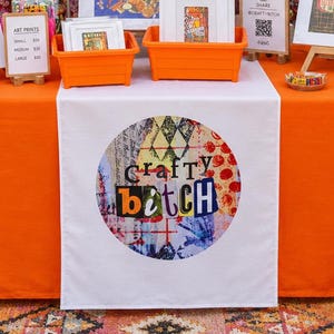 Könnte beinhalten: Ein Verkaufsstand mit einer orangefarbenen Tischdecke und einem kreisförmigen Logo mit der Aufschrift Crafty Bitch. Die Auslage umfasst gerahmte Kunstdrucke, orangefarbene Kunststoffbehälter, eine kleine Topfpflanze und eine Tafel mit dem Text Art Prints Goods Good Vibes.