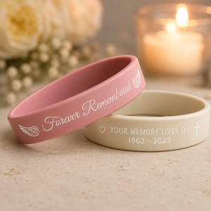 May include: A pink silicone wristband with white text reading Forever Remembered and wing icons, paired with a cream silicone wristband featuring white text reading YOUR MEMORY LIVES ON 1962 - 2023, a heart icon, and a small cross symbol.