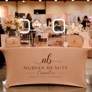 May include: A display table covered in a peach-colored cloth featuring the black text NUBIAN BEAUTY COSMETICS BEAUTY THAT EMPOWERS. The table holds two gold cosmetic bags, clear acrylic organizers filled with makeup brushes, and floral arrangements.