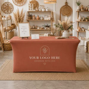 May include: A rectangular terracotta-coloured stretch table cover featuring a white printed logo with an arched botanical design, the text "YOUR LOGO HERE" and "HANDMADE WITH LOVE", and a small heart icon centred on the front panel.