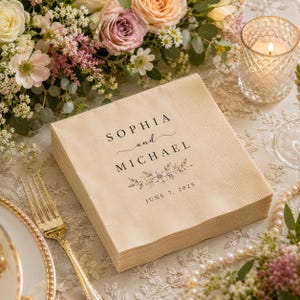 May include: A stack of square beige paper napkins featuring black printed text that reads Sophia and Michael with a floral illustration and the date 7 June 2025. The napkins have a textured border and are placed on a decorative table setting.