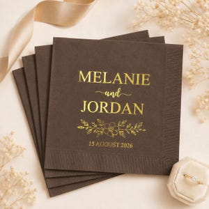 May include: A stack of square dark brown paper napkins featuring gold foil text that reads MELANIE and JORDAN with a floral graphic and the date 15 AUGUST 2026. The napkins have a textured border and are displayed with a gold ribbon and ring.