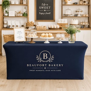 May include: A navy blue fitted tablecloth featuring the white logo of Beaufort Bakery with a central letter B inside a circular laurel wreath, accompanied by the text Sweet Moments, Made With Care, displayed on a rectangular table.