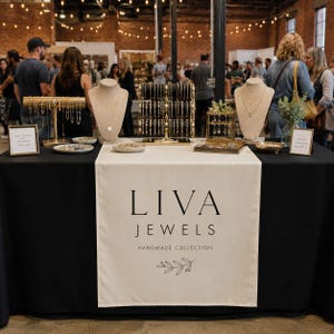 May include: A black table covered with a cream-colored runner featuring the text LIVA JEWELS HANDMADE COLLECTION and a leaf graphic. The table displays jewelry on two beige necklace busts, a gold multi-tier bracelet stand, and a gold jewelry tray.