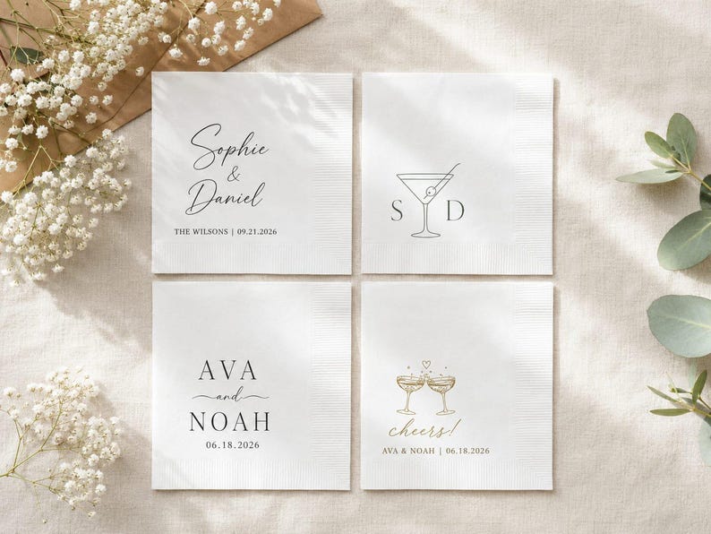 May include: Four square white paper napkins featuring custom black and gold printed designs. The napkins display names like Sophie & Daniel and Ava and Noah, along with dates, martini glass graphics, and the text cheers with a small heart icon.