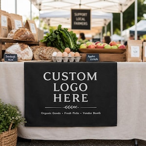 May include: A black table runner with the white text CUSTOM LOGO HERE and the words Organic Goods, Fresh Picks, Vendor Booth. The runner is displayed on a table featuring sourdough bread, a carton of eggs, and a wooden bowl of red and green apples.