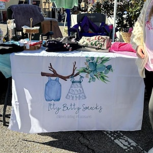 May include: A white tablecloth with the text "Itty Bitty Smocks" and illustrations of children's clothing. The table displays various children's apparel, including a blue denim overall and a white dress. The table is set up at an outdoor market.