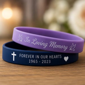 Könnte beinhalten: Zwei Silikonarmbänder, eines in Lila und eines in Marineblau. Das lila Band trägt die Aufschrift "In Loving Memory" mit einer Taubengrafik. Das marineblaue Band hat ein Kreuz, den Text "FOREVER IN OUR HEARTS 1965 - 2023" und eine Herzgrafik.