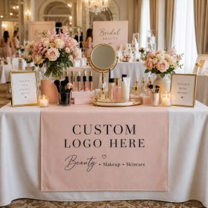 以下が含まれることがあります： CUSTOM LOGO HERE Beauty Makeup Skincareという文字が入ったピンクのテーブルランナーが敷かれた展示テーブル。テーブルの上には、フラワーアレンジメント、ゴールドの鏡、メイクブラシ、スキンケアボトル、そしてEnhance Your Natural BeautyとExclusive Bridal Specialsと書かれた2つの看板が置かれています。