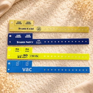 Op de afbeelding: Vier plastic polsbandjes met afneembare lipjes. De bovenste is goudkleurig met 'Team Gold' en 'ONE DRINK'. De tweede is marineblauw met 'Team Navy' en 'ONE DRINK'. De derde is neongeel met 'ALLIANZ 30' en 'I'M IN'. De onderste is blauw met 'vac'.