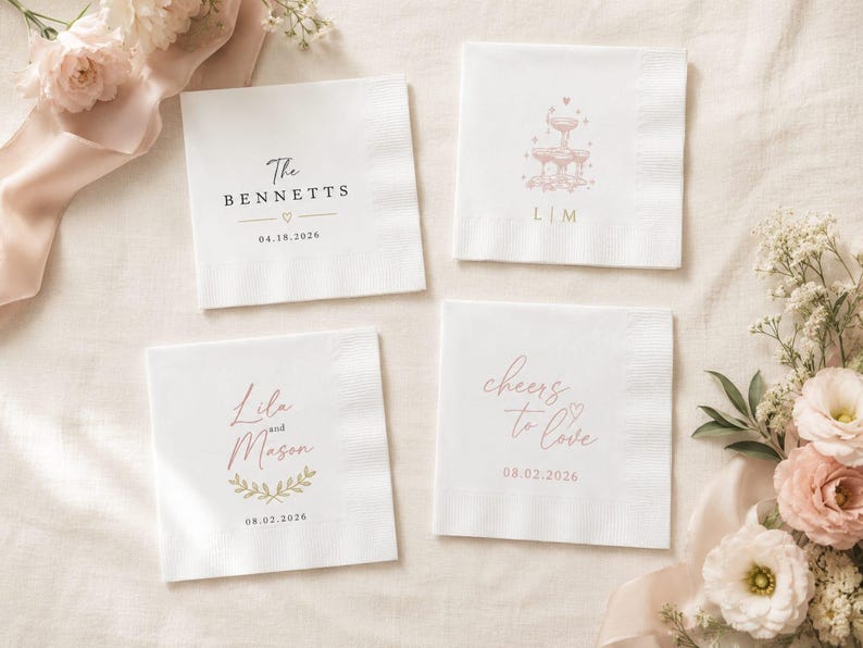 May include: Four square white paper napkins featuring custom printed designs. One displays The Bennetts with 18.04.2026, another a fountain with L|M, one shows Lila and Mason with 02.08.2026 and a leaf motif, and the last reads cheers to love with 02.08.2026.