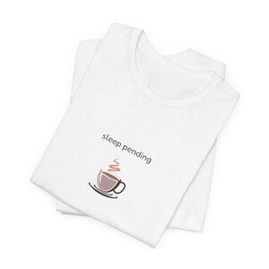 Pode incluir: Uma t-shirt branca com o texto "sleep pending" impresso numa fonte sem serifa castanho-escura acima de um gráfico de uma chávena de café castanha num pires com um pequeno remoinho de vapor vermelho claro a subir da chávena.