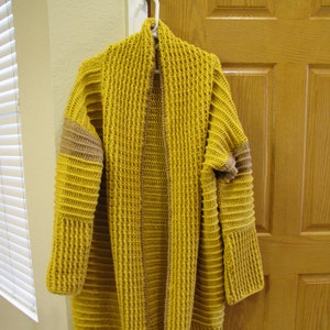 May include: A long, mustard yellow cardigan with brown trim on the sleeves and down the front. The cardigan is crocheted and has a relaxed fit.