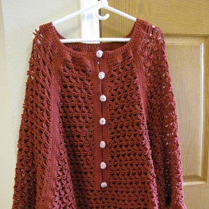 Puede incluir: Un poncho de ganchillo con cuerpo marrón y ribete de ganchillo rosa. El poncho tiene un cierre de botones en la parte delantera.