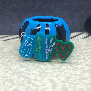 May include: A teal keychain featuring a salt shaker, rosemary, lavender, and a green heart with a red outline. The keychain is in front of a blue and black painted ceramic pot.