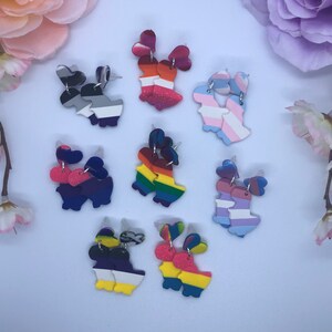 May include: A collection of handmade polymer clay earrings in various pride flag designs. Each pair features a heart-shaped top and a stylized figure, with colors representing different LGBTQ+ identities. The earrings are displayed on a white surface.