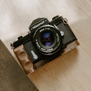 May include: A black Nikon Nikomat camera with a brown leather grip and a 50mm f/1.8 lens.