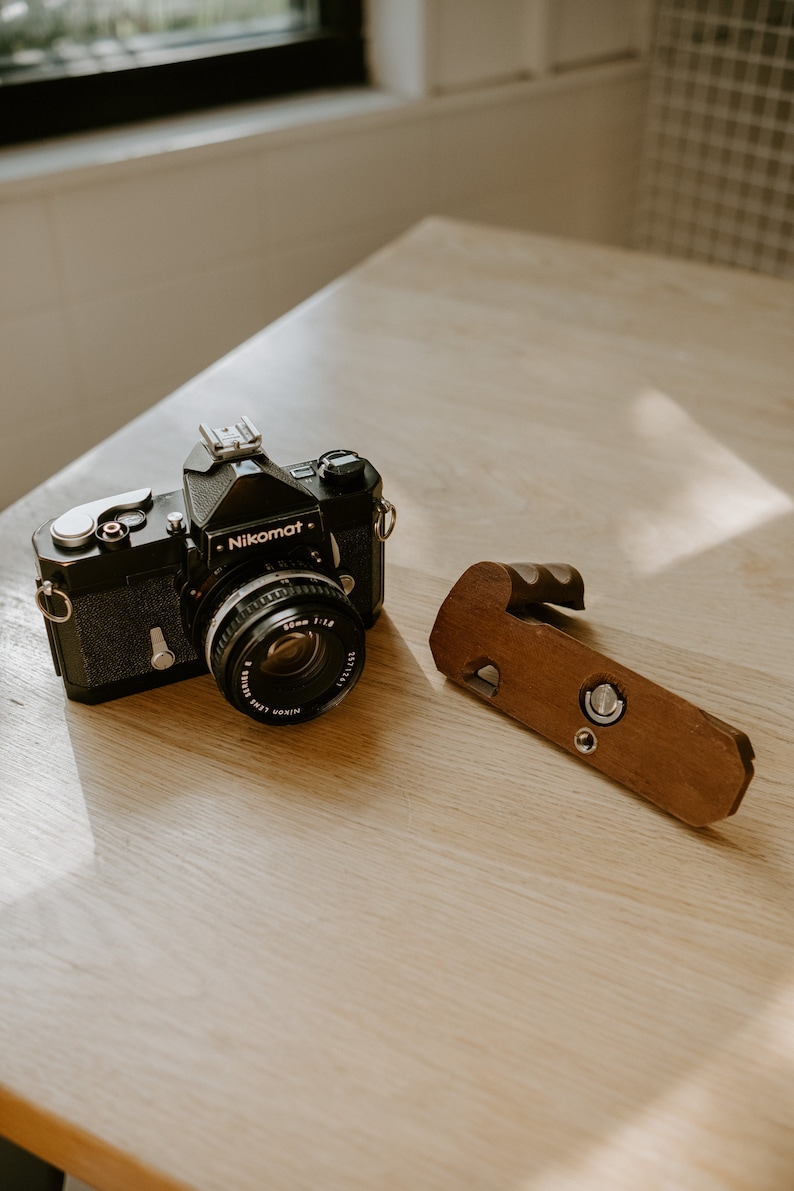 May include: A black Nikon Nikomat camera with a 50mm lens and a wooden hand grip.