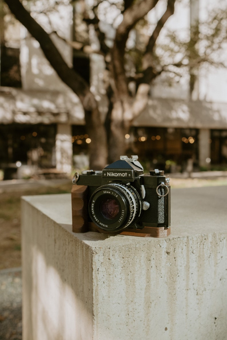 May include: A black Nikon Nikomat camera with a 50mm 1:1.8 lens, sitting on a concrete surface.