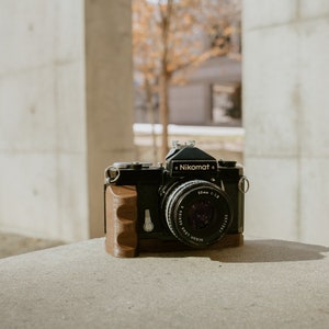 May include: A black Nikon Nikomat camera with a brown leather grip and a 50mm f/1.8 lens. The camera is sitting on a concrete surface.