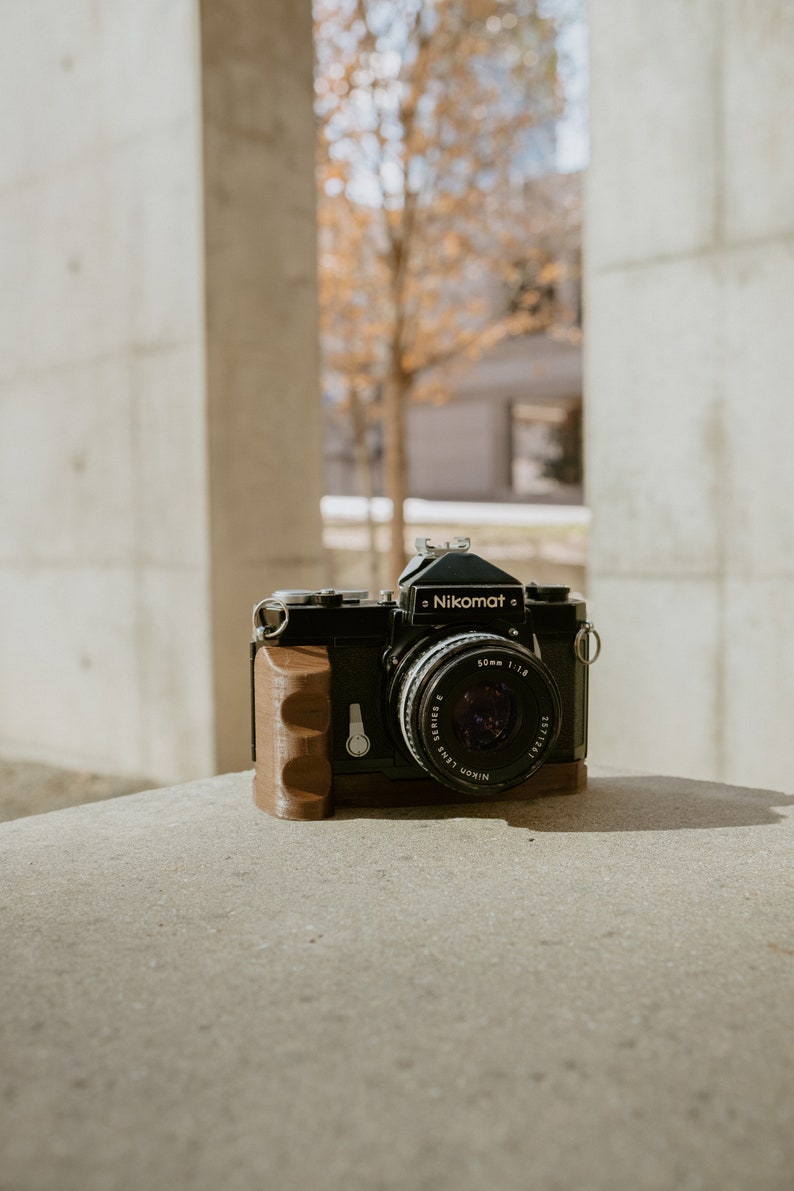 May include: A black Nikon Nikomat camera with a brown leather grip and a 50mm f/1.8 lens. The camera is sitting on a concrete surface.