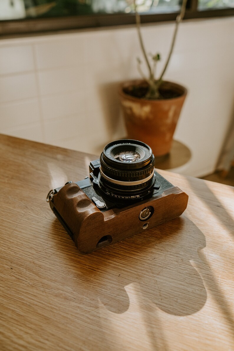 May include: A black camera with a wooden grip and a silver lens. The camera is sitting on a wooden table.