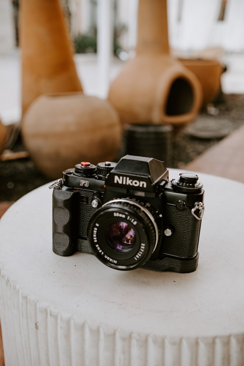 Puede incluir: Una c&aacute;mara de pel&iacute;cula Nikon F3 negra con una lente de 50 mm. La c&aacute;mara est&aacute; sobre una superficie de hormig&oacute;n blanco.