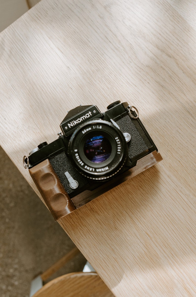 May include: A black Nikon Nikomat camera with a brown leather grip and a 50mm f/1.8 lens.