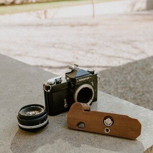 May include: A black Nikon Nikomat camera with a lens attached, a separate lens, and a wooden camera grip.