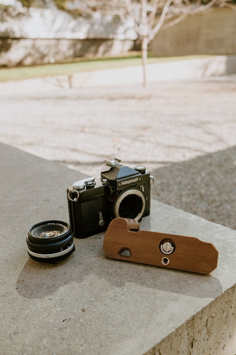 May include: A black Nikon Nikomat camera with a lens attached, a separate lens, and a wooden camera grip.