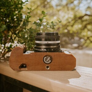 May include: A vintage camera with a wooden body and a black lens. The camera is sitting on a wooden surface.