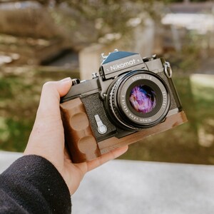 May include: A black Nikon Nikomat camera with a brown leather grip and a 50mm f/1.8 lens.