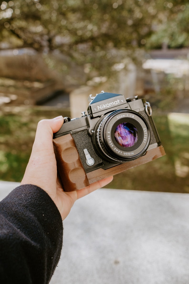 May include: A black Nikon Nikomat camera with a brown leather grip and a 50mm f/1.8 lens.