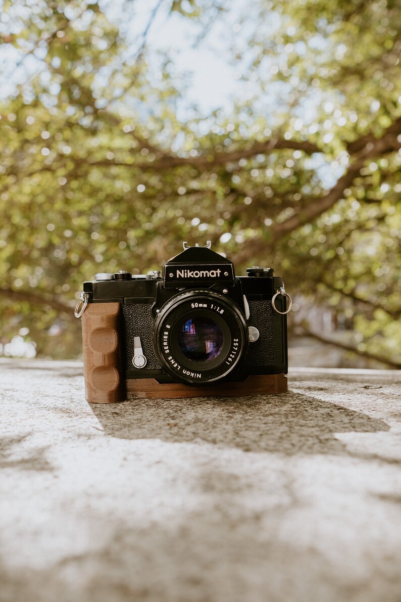 May include: A black Nikon Nikomat camera with a 50mm f/1.8 lens. The camera has a brown leather grip and a silver strap.