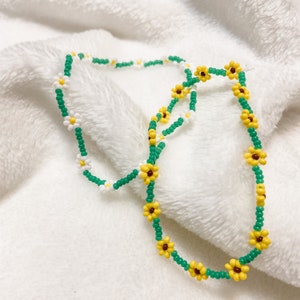 May include: A green and yellow beaded necklace with white and yellow flower accents. The necklace is made of small beads and features a repeating pattern of flowers.