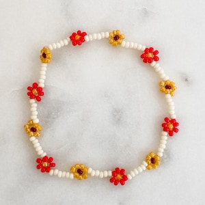 May include: A white beaded bracelet with red and yellow flower charms. The bracelet is made of small white beads and features alternating red and yellow flower charms.